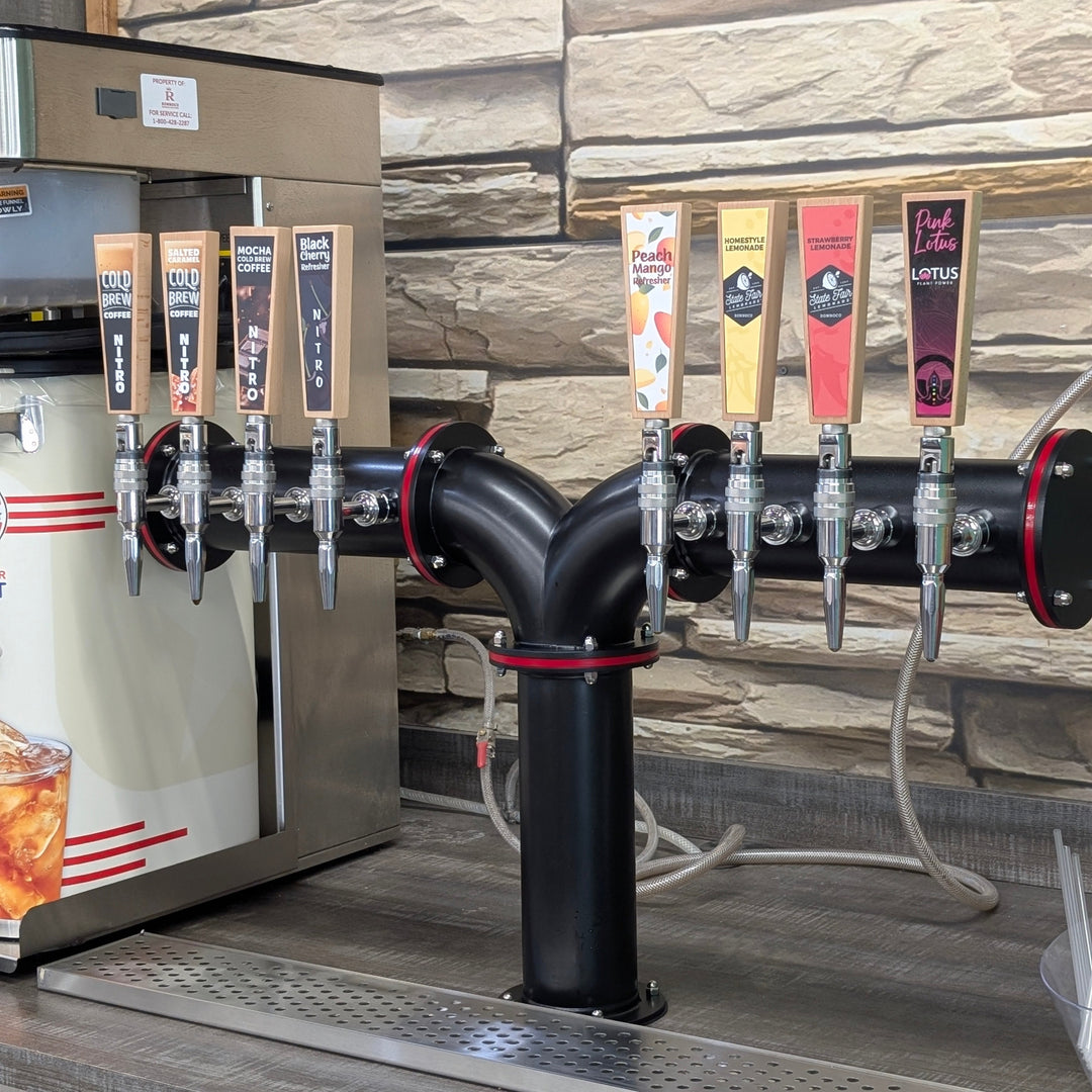 A commercial beverage tap system with multiple beer taps mounted on a black Y-pipe, set against a stone-tile wall and next to a stainless steel dispenser.